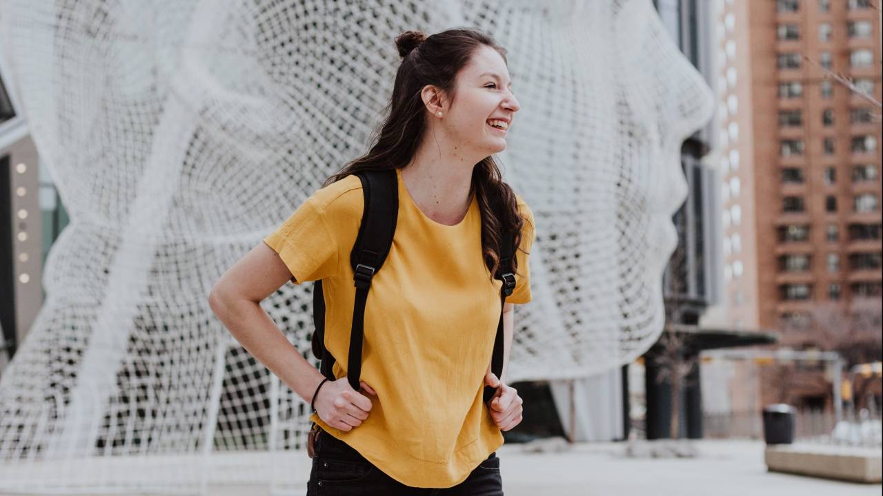 Uma menina de cabelos escuros e longos está em um local aberto, ela veste uma camiseta amarela e carrega uma mochila nas costas. Olhando para o lado, ela sorri