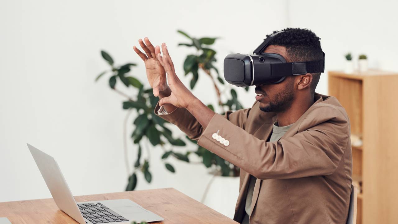 Um homem vestido de terno marrom está sentado em frente a um computador. Ele está com um óculos de realidade virtual no rosto enquanto faz gestos com as mãos