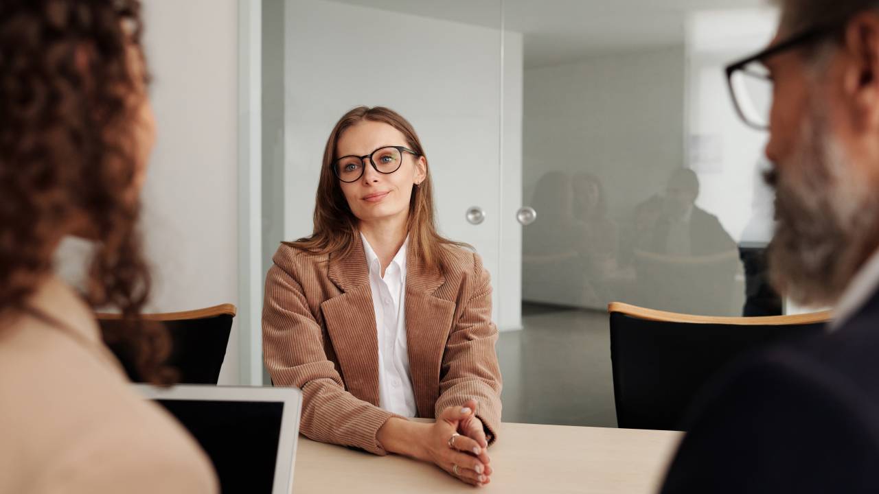 Mulher com óculos de grau, blazer marrom e camisa branca está em uma entrevista de emprego