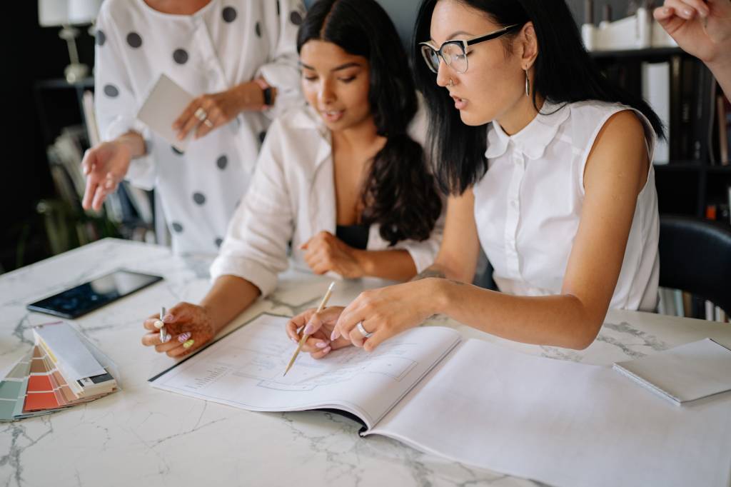 Três mulheres conversam em uma reunião apontando um caderno de desenho. Uma delas usa óculos gatinho com armacão preta e detalh branco, outra uma blusa de poá e outra uma camisa branca com top preto