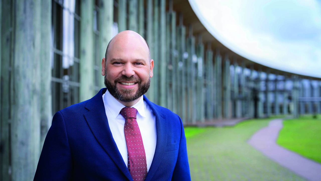 Daniel está em um ambiente externo, vestindo um terno azul, camisa branca e gravata vermelha e sorri