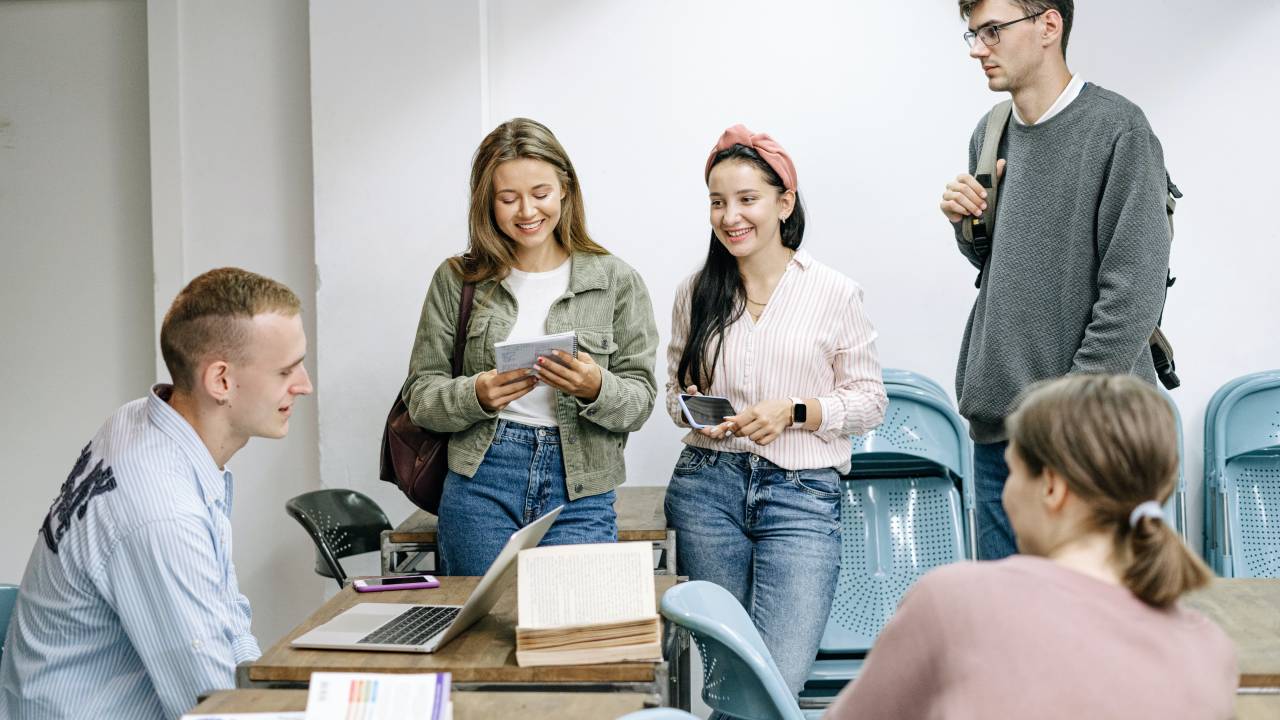 Cinco pessoas estão ao redor de uma mesa. Na mesa tem um computador, livros e cadernos. Três das pessoas estão de pé e as outras duas sentadas