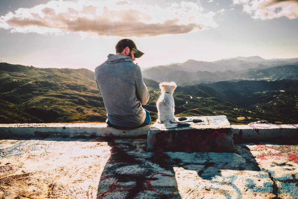 Um homem está no pico de uma montanha ao lado de seu cachorro, que é branco e peludo