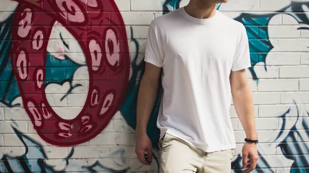 Homem com camiseta branca e calça creme está em frente a um muro com grafite