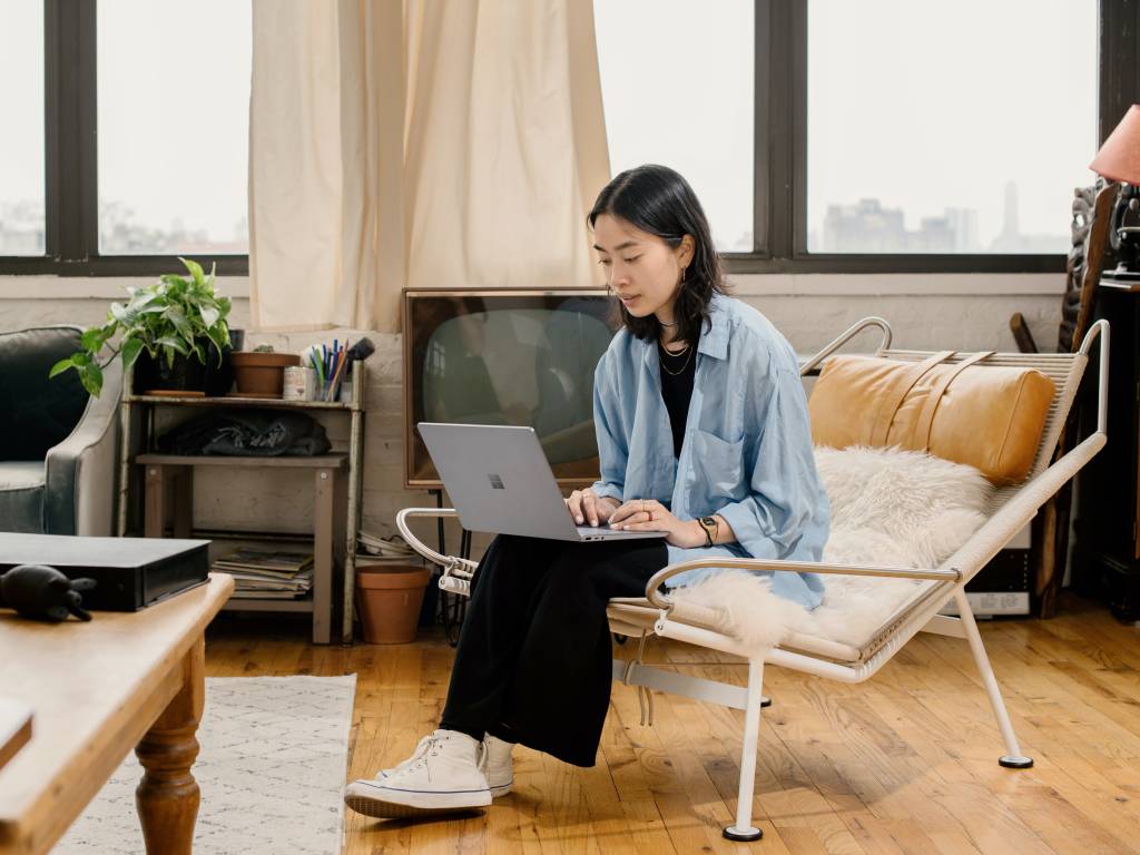 Uma mulher com traços asiáticos está sentada em uma cadeira e tem um computador no colo. Ela veste uma camisa larga azul e calça preta. O ambiente é uma sala de estar