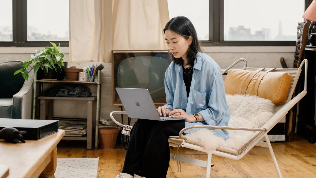 Uma mulher com traços asiáticos está sentada em uma cadeira e tem um computador no colo. Ela veste uma camisa larga azul e calça preta. O ambiente é uma sala de estar