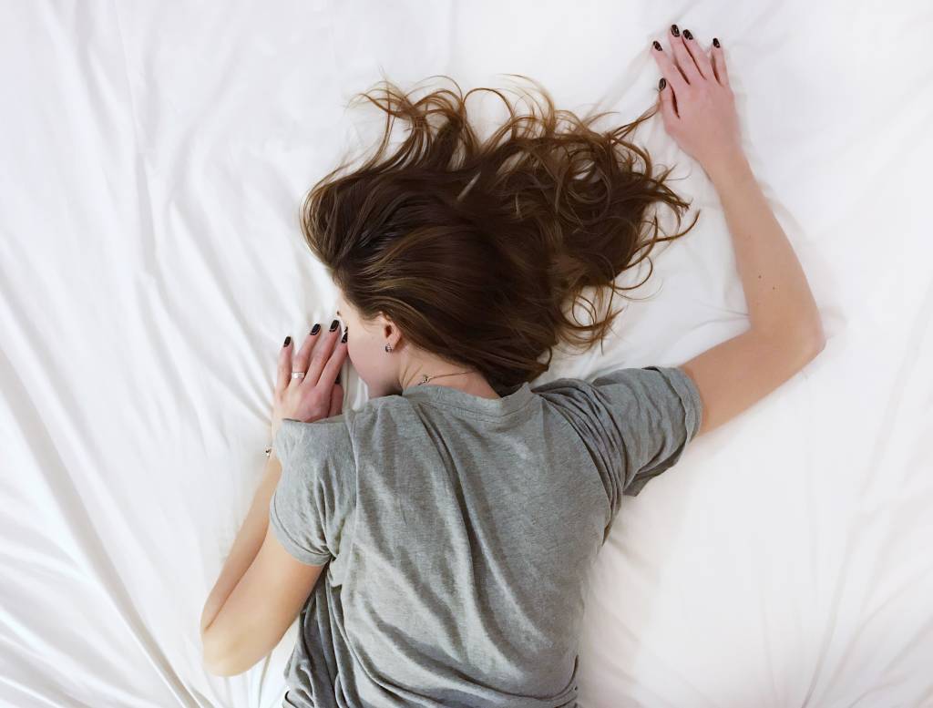 Mulher de camiseta cinza e cabelo liso castanho está deitada em uma cama com lençois brancos