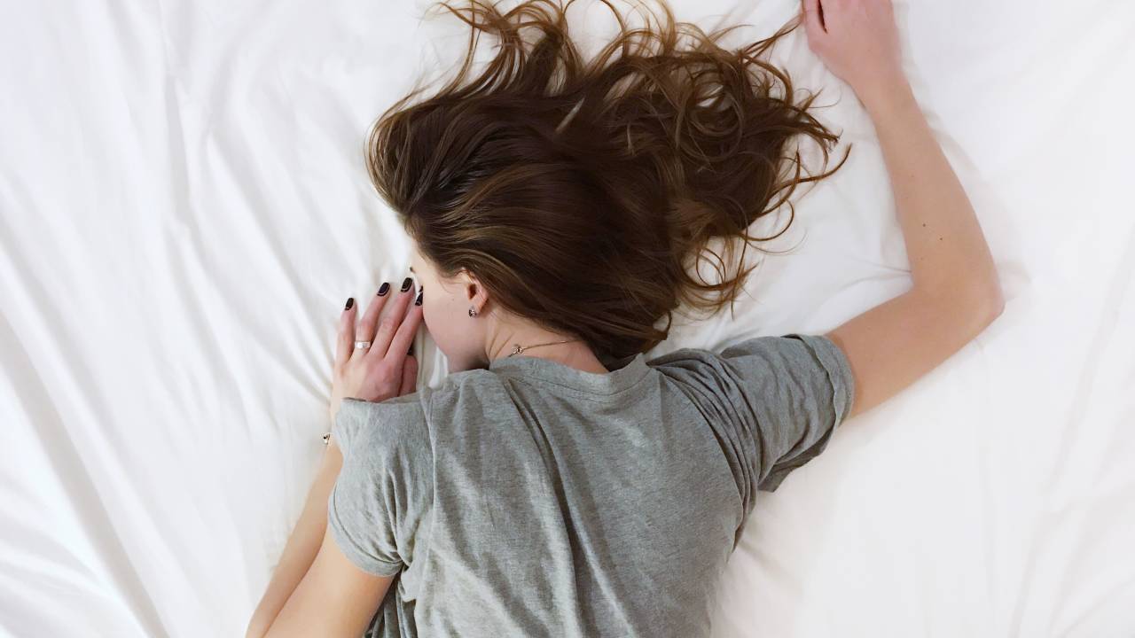 Mulher de camiseta cinza e cabelo liso castanho está deitada em uma cama com lençois brancos