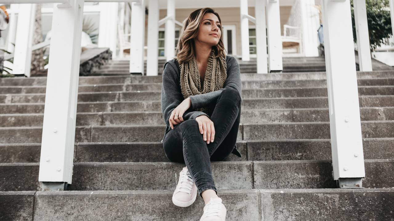 Mulher de jeans e blusa cinza chumbo usa lenço no pescoço. Ela está sentada em uma escadaria contemplando o horizonte