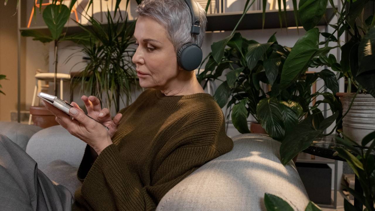 Uma mulher está sentada em um sofá, tem cabelos curtos e grisalhos e usa um fono de ouvido. Ela mexe no celular e, atrás dela, há prateleiras com plantas