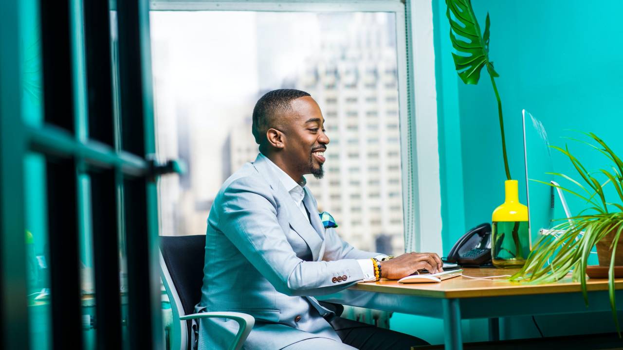 Um homem negro, vestido com um paletó azul claro, está sentado em frente a uma mesa de escritório. O ambiente é bastante colorido e ele sorri para a tela