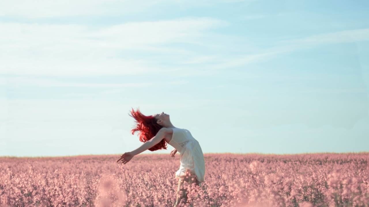 Uma mulher veste um vestido claro e leve, tem os cabelos vermelhos, e está no meio de uma imensidão de flores rosas