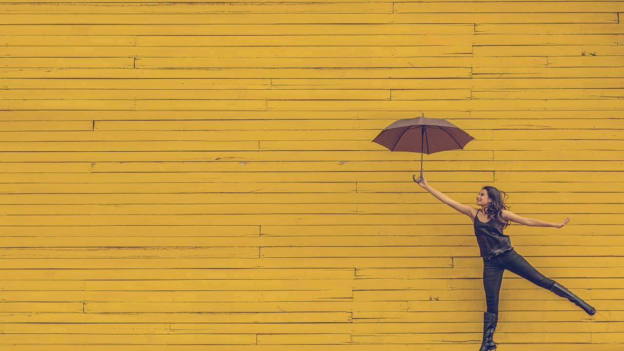Mulher salta com um guarda-chuva em frente a uma parede amarela