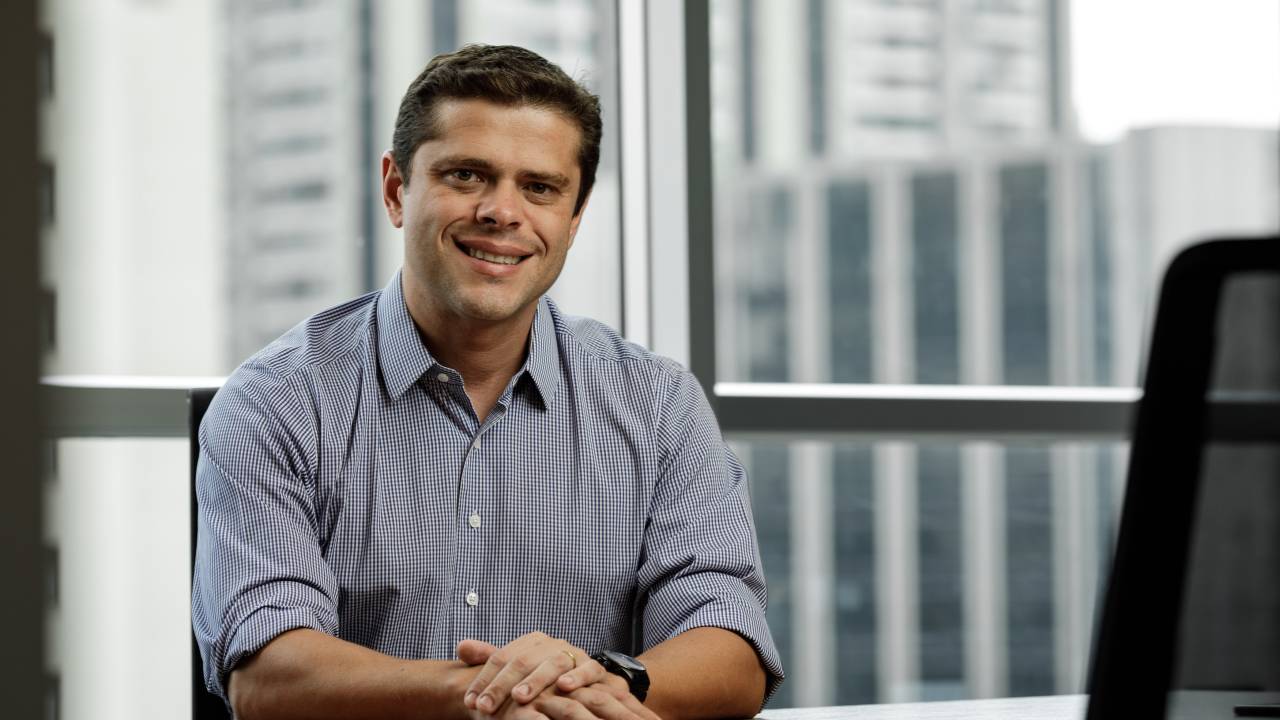 Rodrigo Vianna, CEO da Mappit, está vestindo uma camisa azul, sorrindo e sentado em frente a uma mesa