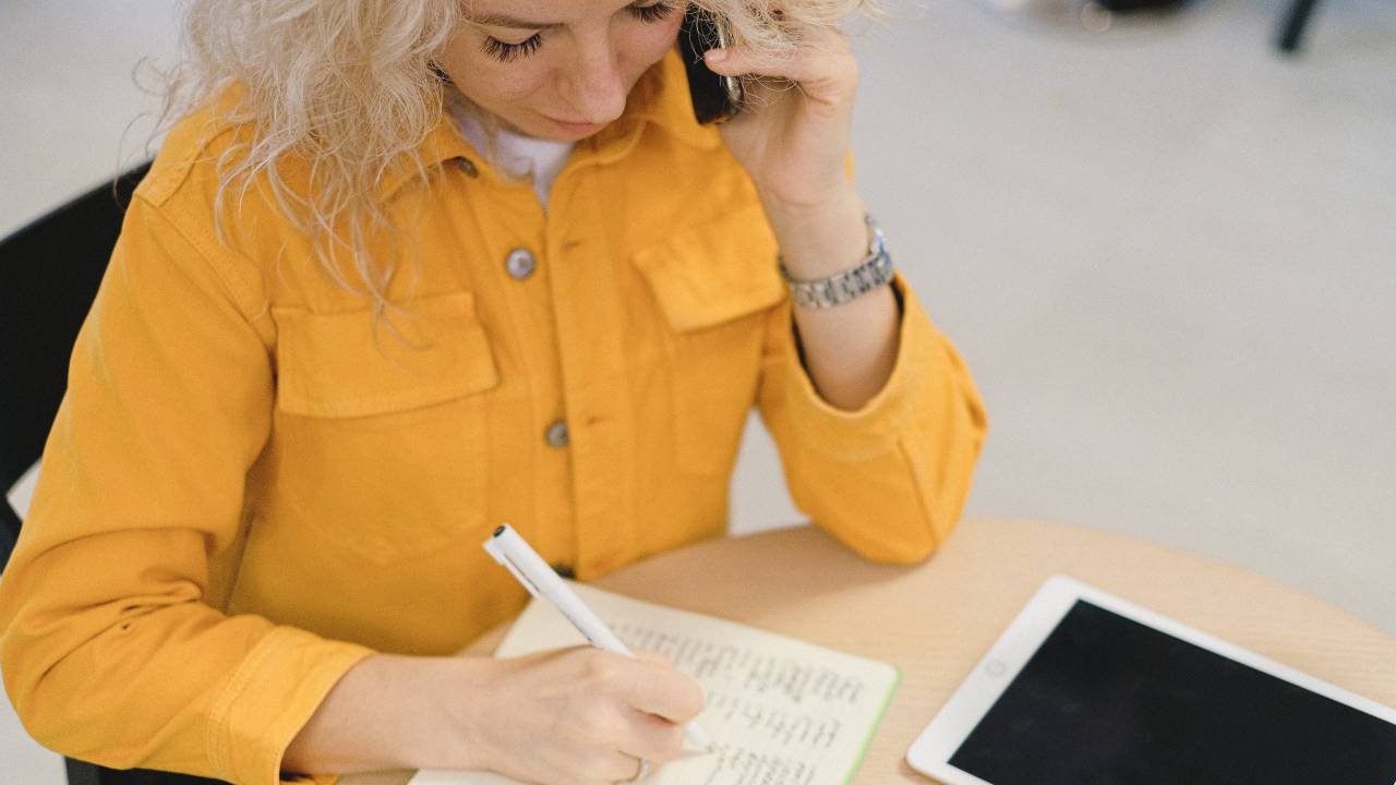 Uma mulher fala ao telefone enquanto faz anotações em um caderno. Ela usa uma camisa amarela e é loira