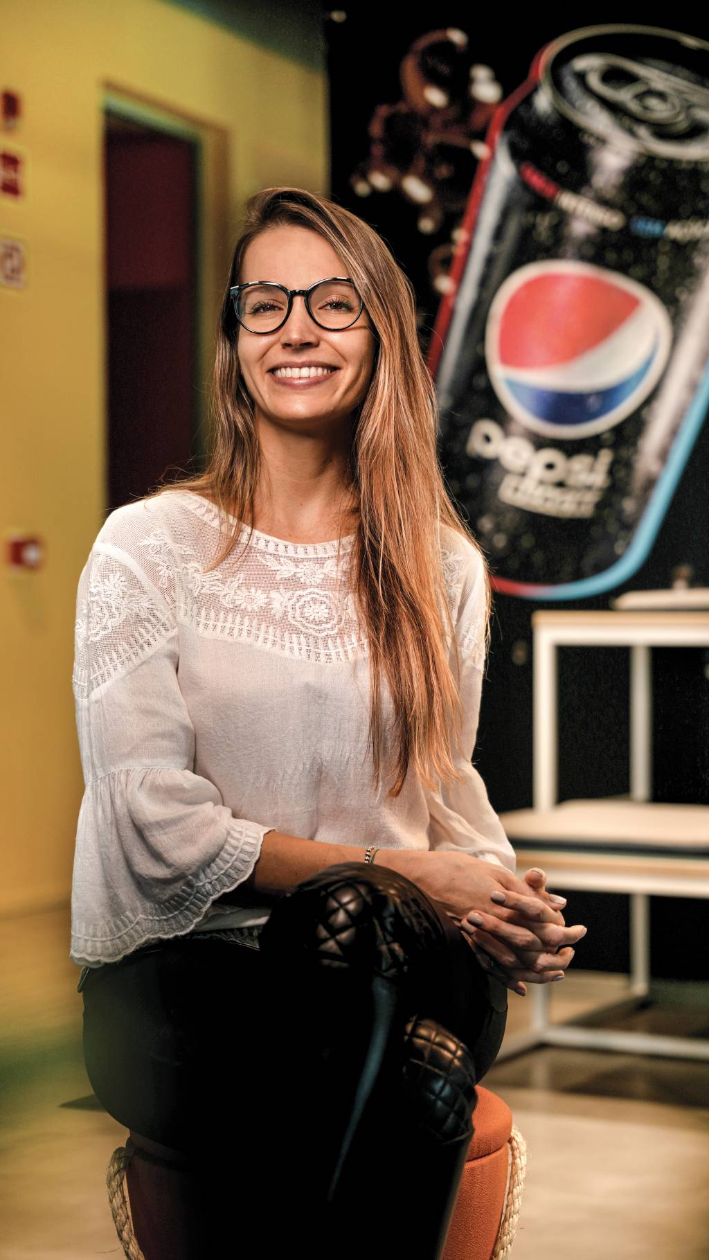 Aline está sentada, usa uma camisa de manga longa branca e uma calça preta. Ela está sorrindo, tem os cabelos longos e está em frente a um banner da Pepsi