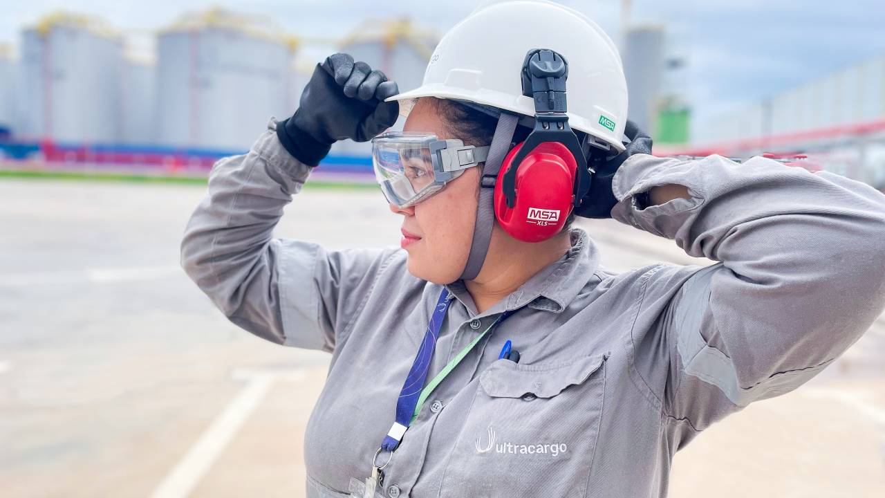 Uma mulher veste um uniforme de operadora, usa óculos de proteção, luvas e um capacete