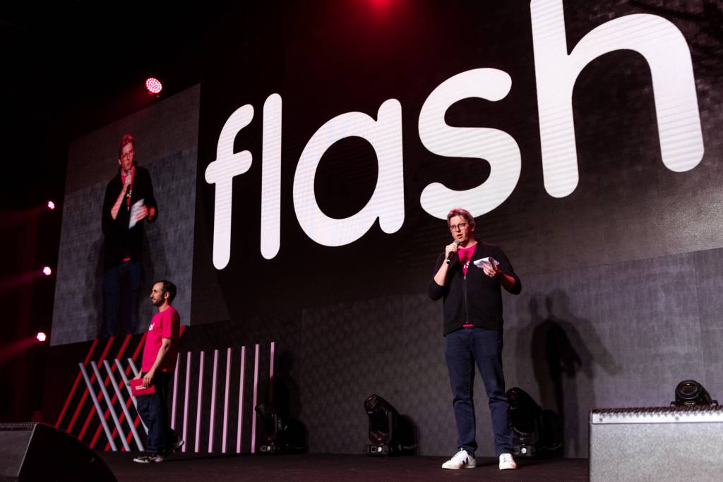 Ricardo e Guilherme estão em um palco com fundo preto e com o logo da Flash em branco