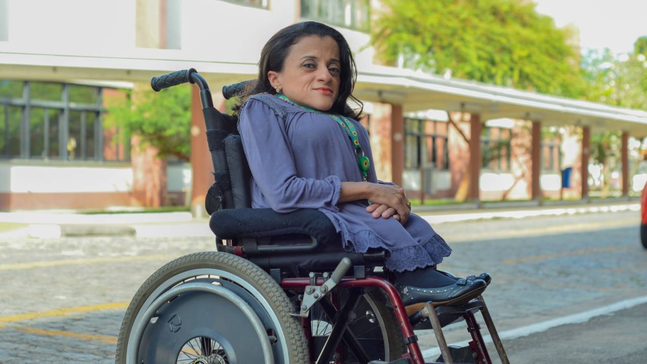 Thais Pessanha está sentada em uma cadeira de rodas. Ela está de lado, olhando para a foto, vestindo roupas em tom de azul em um ambiente externo