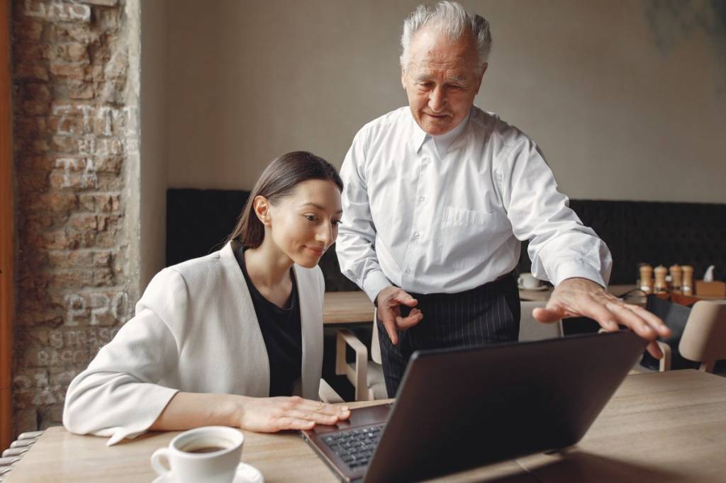 imagem de dois colegas de trabalho, um homem idoso e grisalho ao lado de uma mulher jovem e branca.