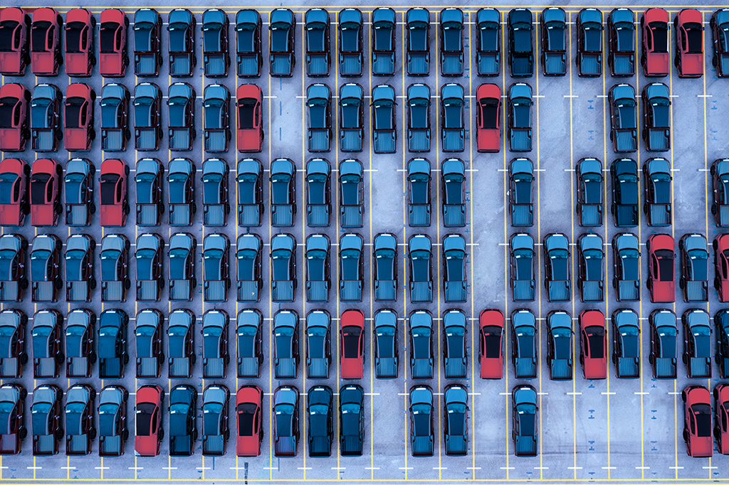 Imagem de vista área de um estacionamento com carros ocupando a maioria das vagas. A predominância é de carros da cor azul. A minoria são carros vermelhos.