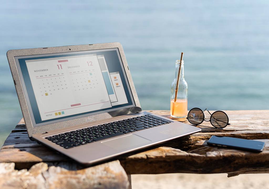 Composição de um laptop, um suco, óculos de sol e celular sobrepostos em uma tábua de madeira. Ao fundo, há um oceano.