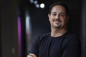 Um homem de cabelos escuros, barba e bigode vestido uma camiseta preta. Ele est de braos cruzados.