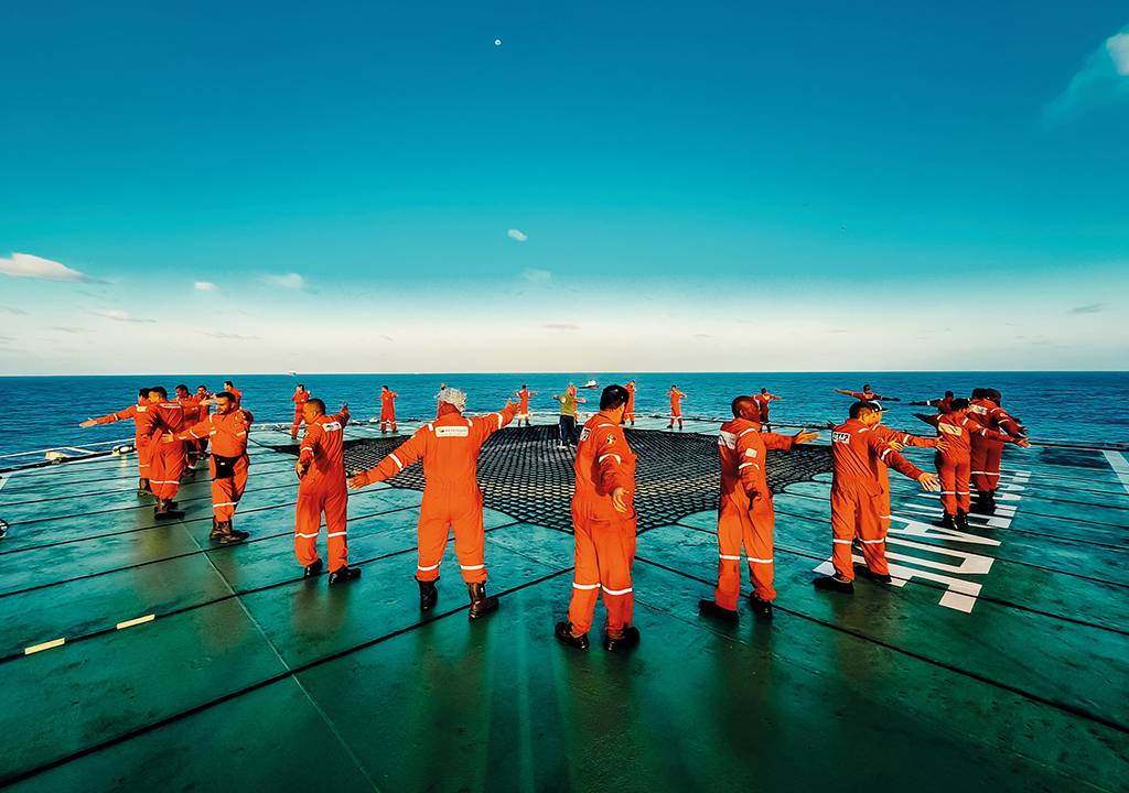Vários homens com uniformes laranjas em cima de uma plataforma de petróleo. Ao fundo está o oceano.