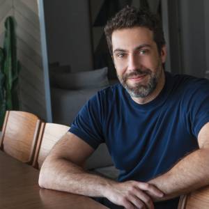 Foto de um homem de barba sorrindo vestindo camiseta azul, sentado a mesa.