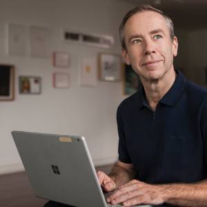 Foto de um homem sorrindo, vestindo camisa azul marinho, usando um notebook numa sala decorada com quadros.