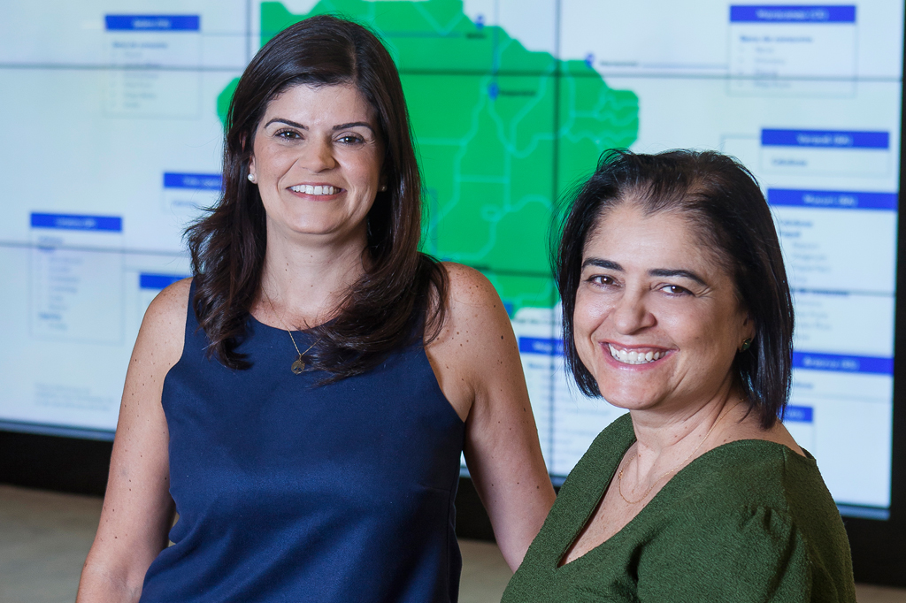 VCRH_95_IA_no_RH_4 Foto de duas mulheres sorrindo, uma veste vestido azul e outra veste camisa verde. Ao fundo, um grande monitor com o mapa do Brasil.
