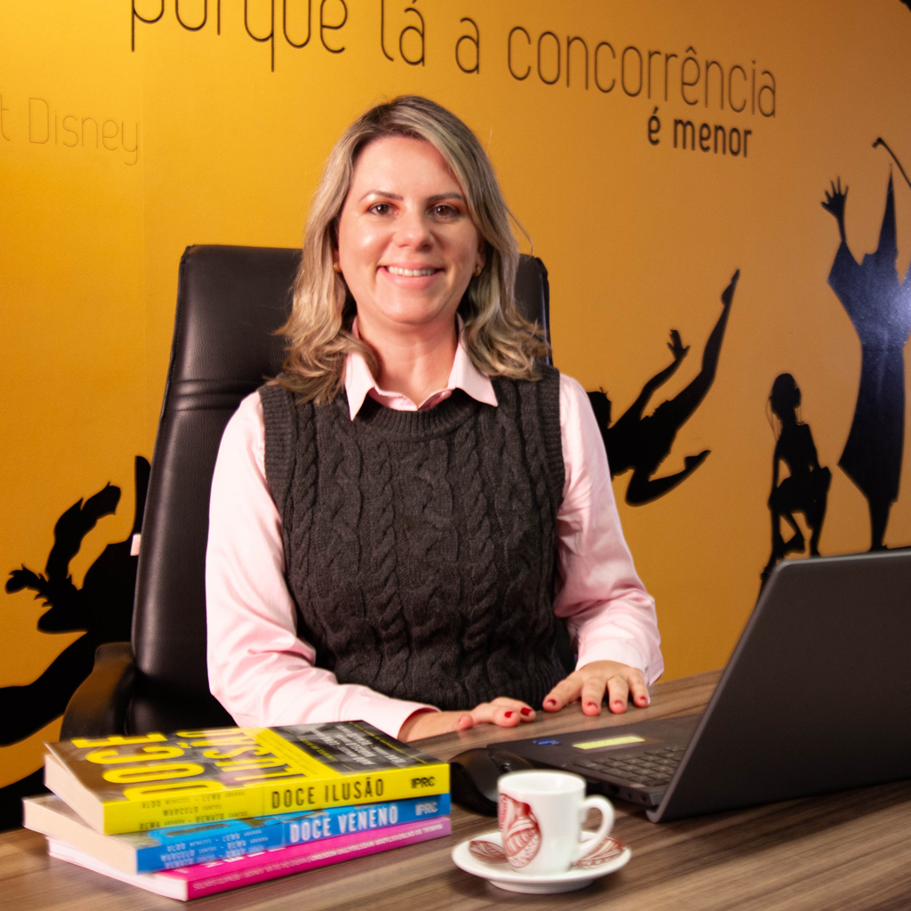 Imagem de uma executiva sorrindo, sentada diante de um notebook, vestindo uma camisa rosa e um colete preto.