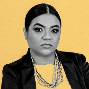 Imagem, em fundo amarelo, de uma mulher com semblante sério, vestindo terno preto e colar.