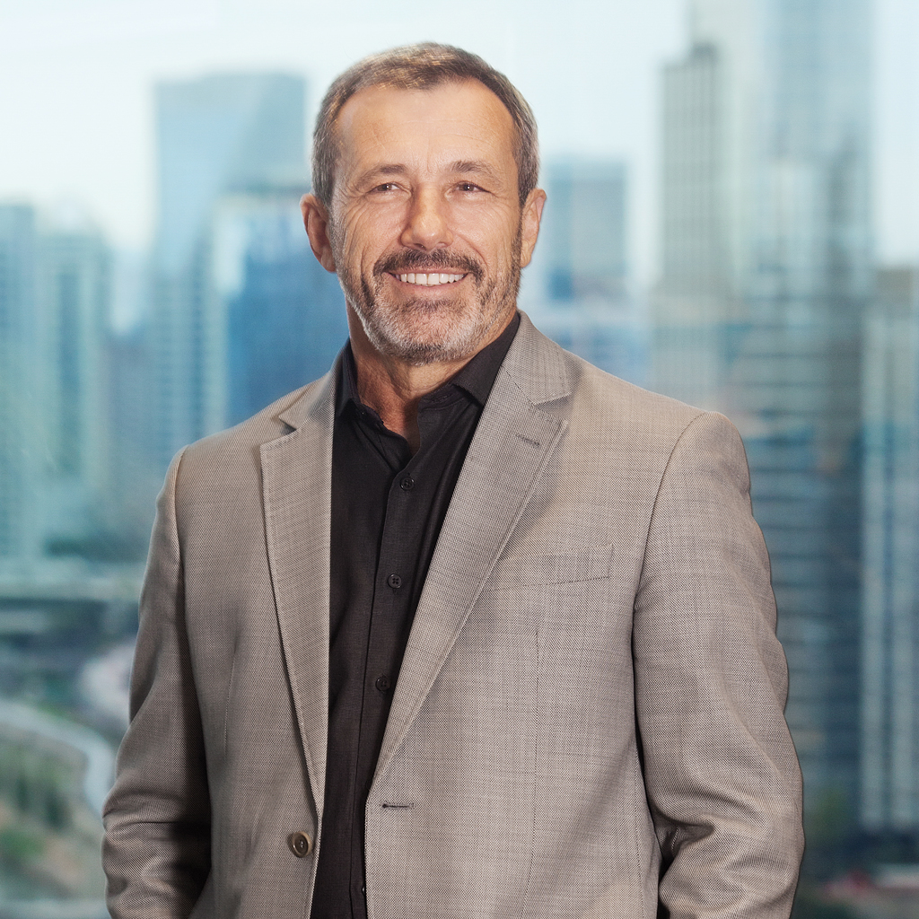 VCRH_97_Olhar_de_Presidente_1 Imagem de um homem sorrindo, vestindo terno cinza e camisa preta. Ao fundo, uma grande janela de vidro transparecendo outros prédios da cidade.