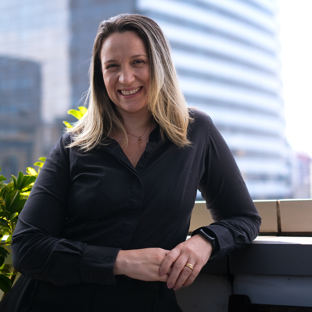 Imagem de uma mulher sorrindo, de cabelos loiros, vestindo uma roupa preta. Ao fundo, vê-se um grande edifício.