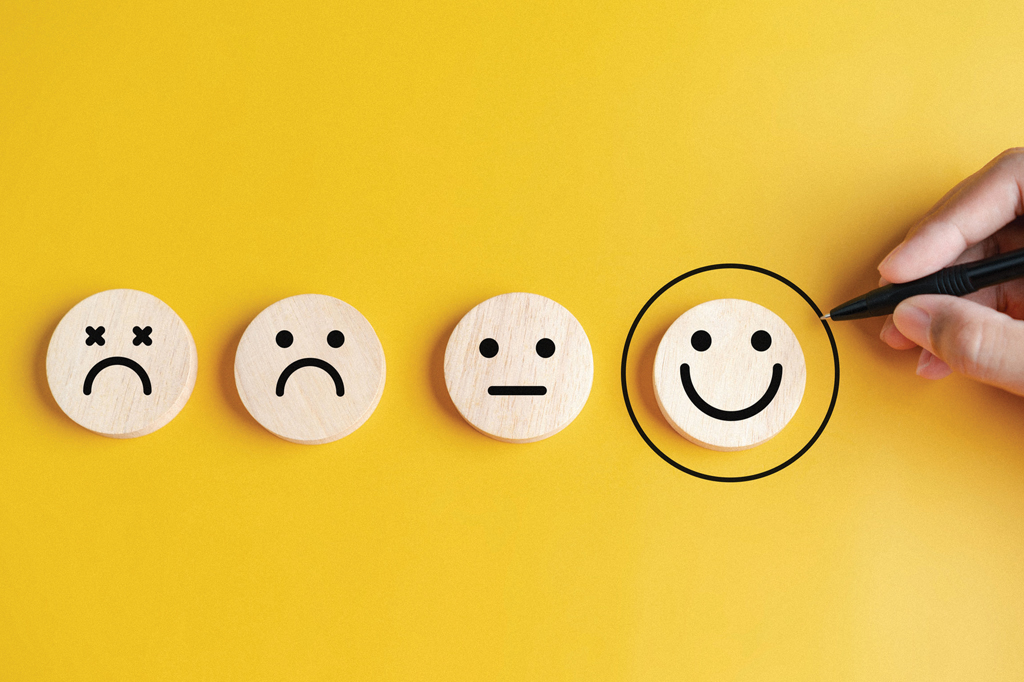Imagem, em fundo amarelo, com emojis representando quatro tipos de estado de espírito – de triste a feliz. Uma caneta circula o emoji de rosto do sorriso feliz.