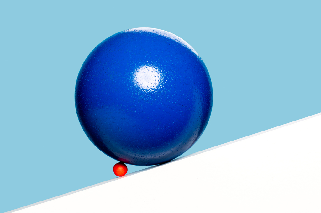 Imagem, em fundo azul claro, de uma pequena bola vermelha bloqueando uma bola azul muito maior em uma rampa branca.