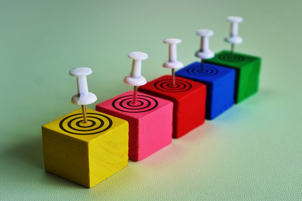 Imagem, em fundo verde, de cinco cubos coloridos. Cada um deles tem no topo um alvo com um alfinete espetado.