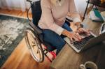 Uma mulher sentada em uma cadeira de rodas digitando em um notebook apoiado em uma mesa.