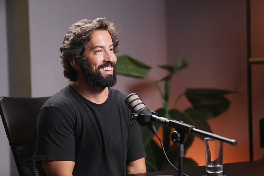 Um homem sorrindo de cabelo ondulado e barba vestindo uma camiseta preta.