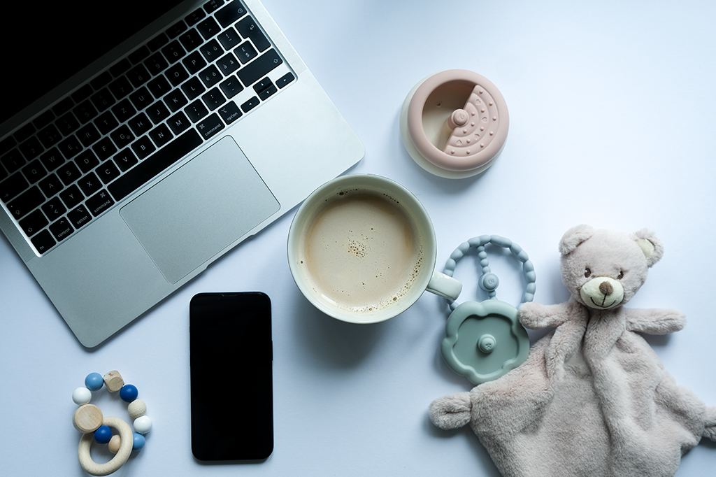 Composição com notebook ao lado de acessórios de criança, como pelúcia e chocalho. Ao centro, uma xícara de café.