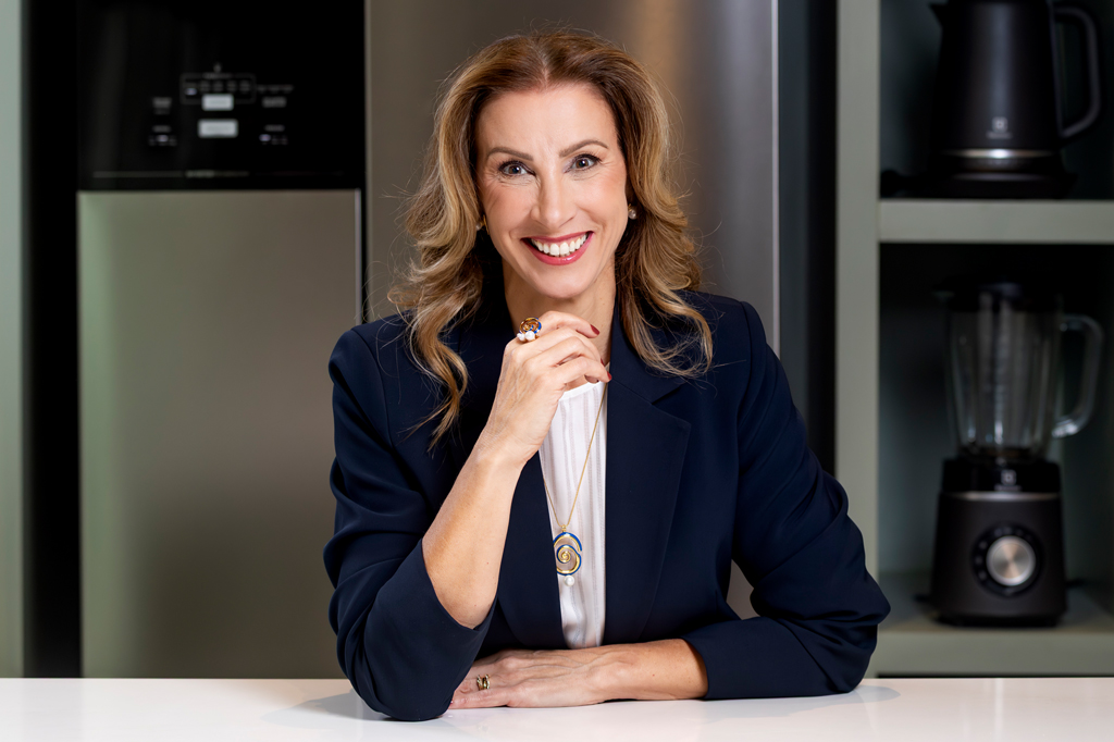Imagem de uma mulher sorrindo, de cabelos loiros, vestindo um elegante casaco azul marinho. Ao fundo, vê-se uma estante com eletrodomésticos e uma geladeira.