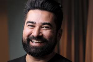VCRH_99_IA_Coach_1 Imagem de um homem, de cabelos e barba preta, sorrindo. Ao fundo vê-se uma cortina marrom.