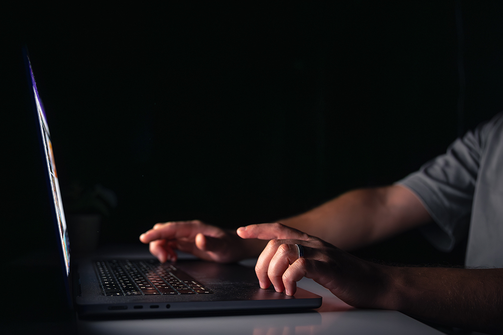 Mãos de um homem trabalhando e digitando no teclado de um notebook à noite.