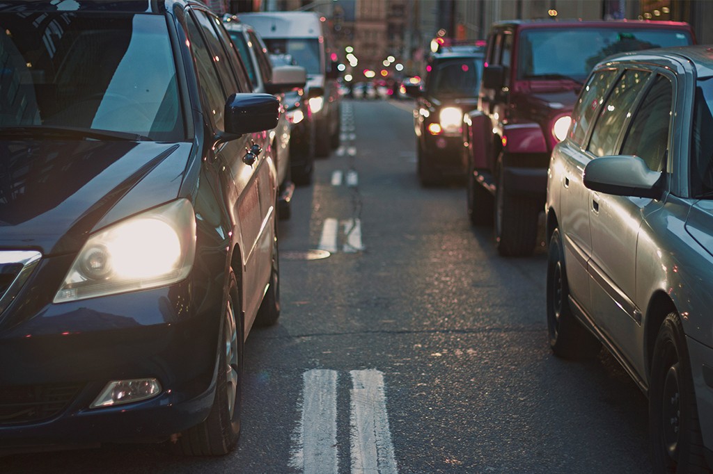 Carros parados no trânsito em uma rua urbana, com faróis acesos e luzes de freio ao fundo, indicando congestionamento ao entardecer