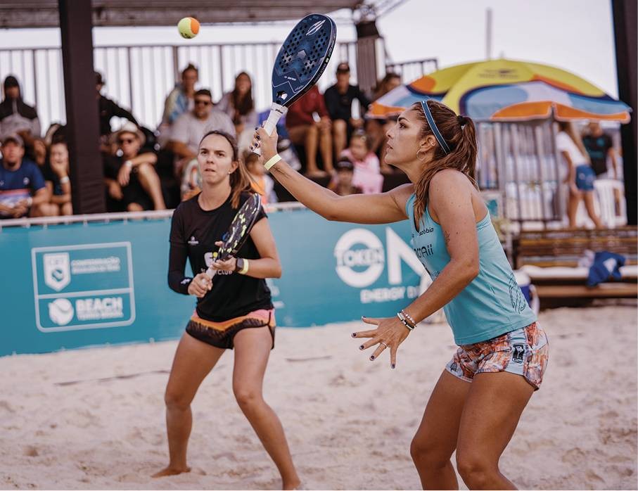 Duas mulheres jogando beach tennis na areia. Uma, de regata azul e shorts estampados, rebate a bola amarela com a raquete. A outra, de camiseta preta e shorts coloridos, observa atenta. Pessoas assistem ao fundo, sob um guarda-sol listrado