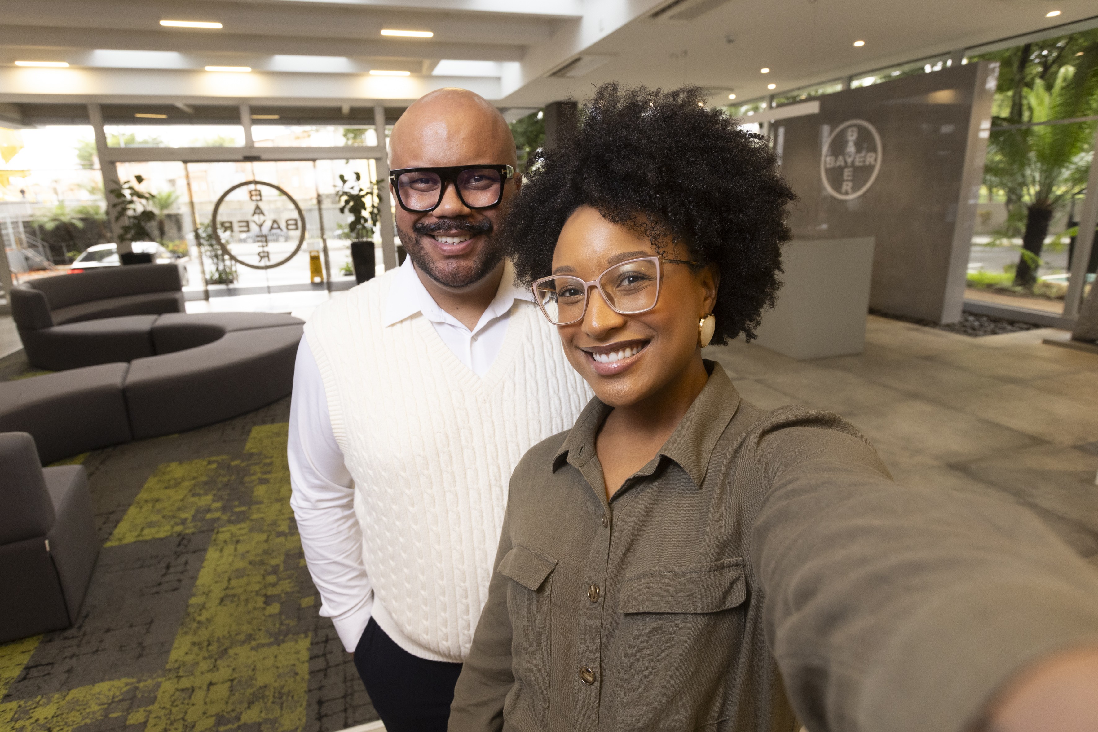 Um homem careca de óculos escuros e um colete de tricô creme sorri ao lado de uma mulher de cabelo crespo e óculos claros, vestindo uma camisa verde-oliva. Ambos sorriem para a câmera em um ambiente corporativo moderno com o logotipo da Bayer visível ao fundo